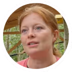 Close-up of a woman with light skin and red hair speaking outdoors with greenery in the background.